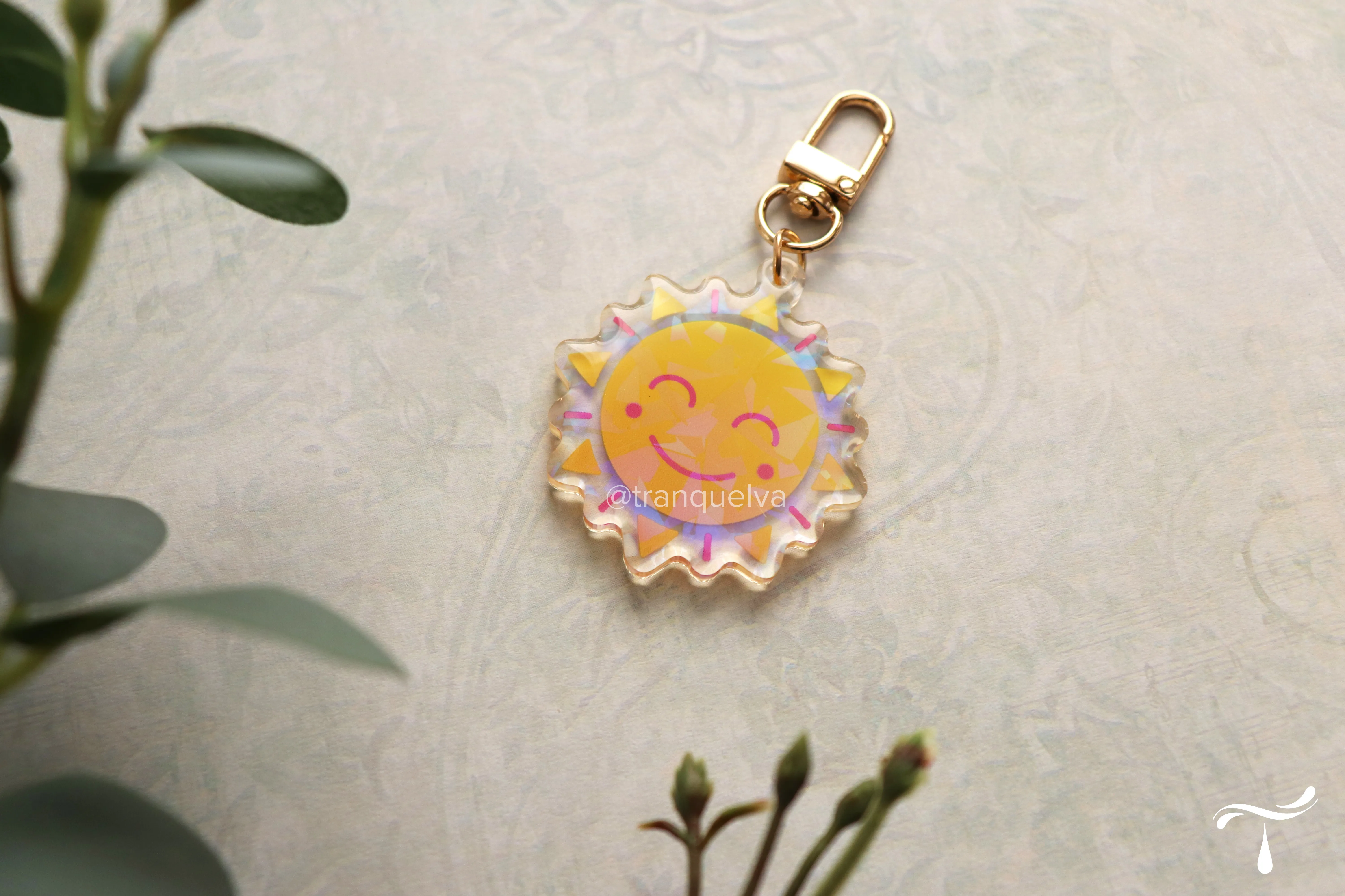 A glittery acrylic keychain of a smiling sun with a gold clasp, photographed beside eucalyptus on a soft floral background