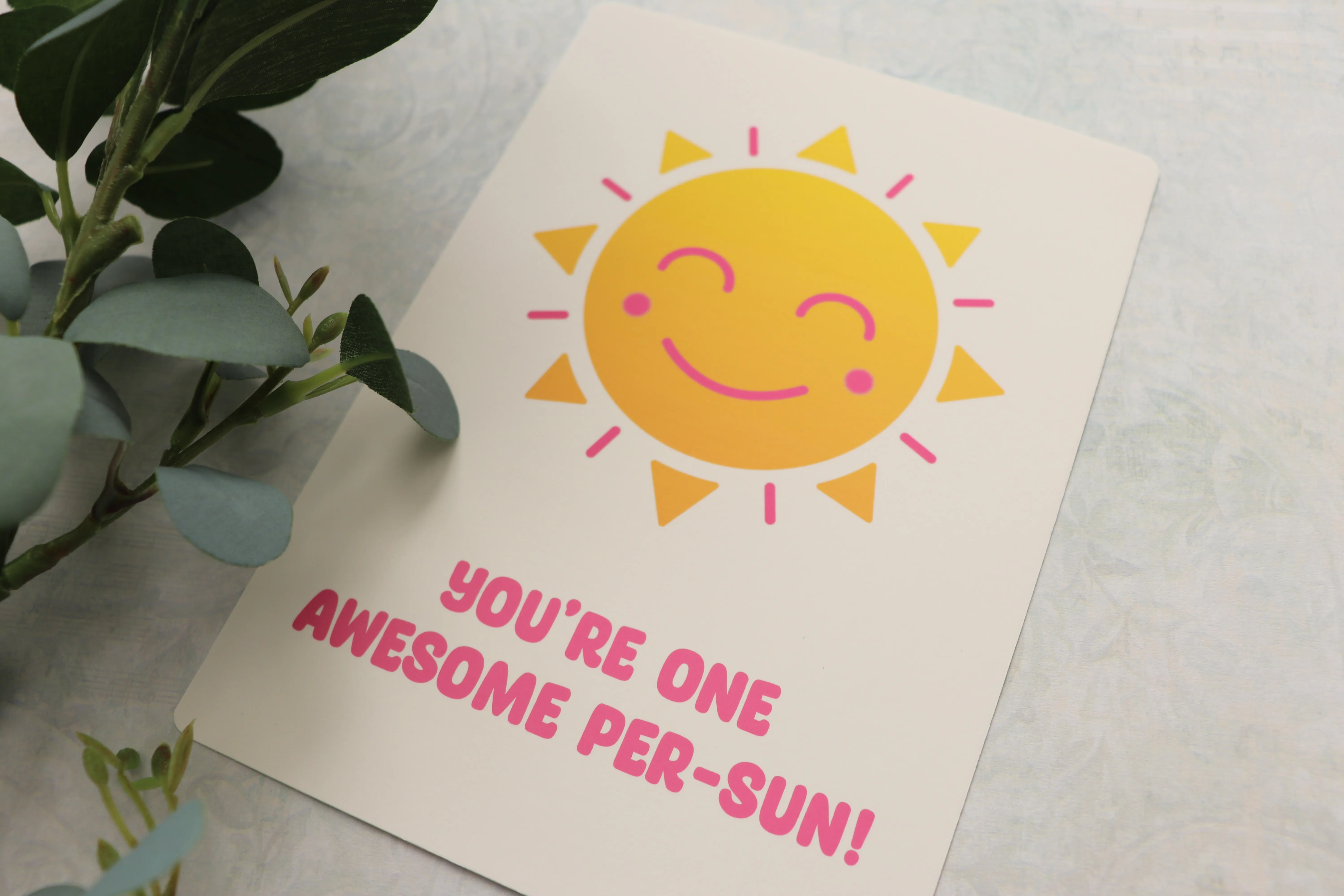 Art print of a smiling yellow sun with the words You're one awesome per-sun in pink, beside eucalyptus